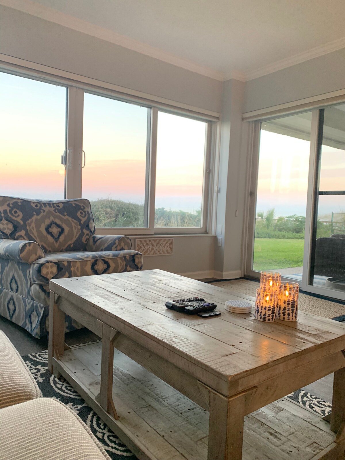 A comfortable living space features a light-colored sofa and an accent chair with a geometric pattern. A rustic wooden coffee table sits in the center, adorned with candles and a remote. Large windows frame scenic views of the ocean at sunset.
