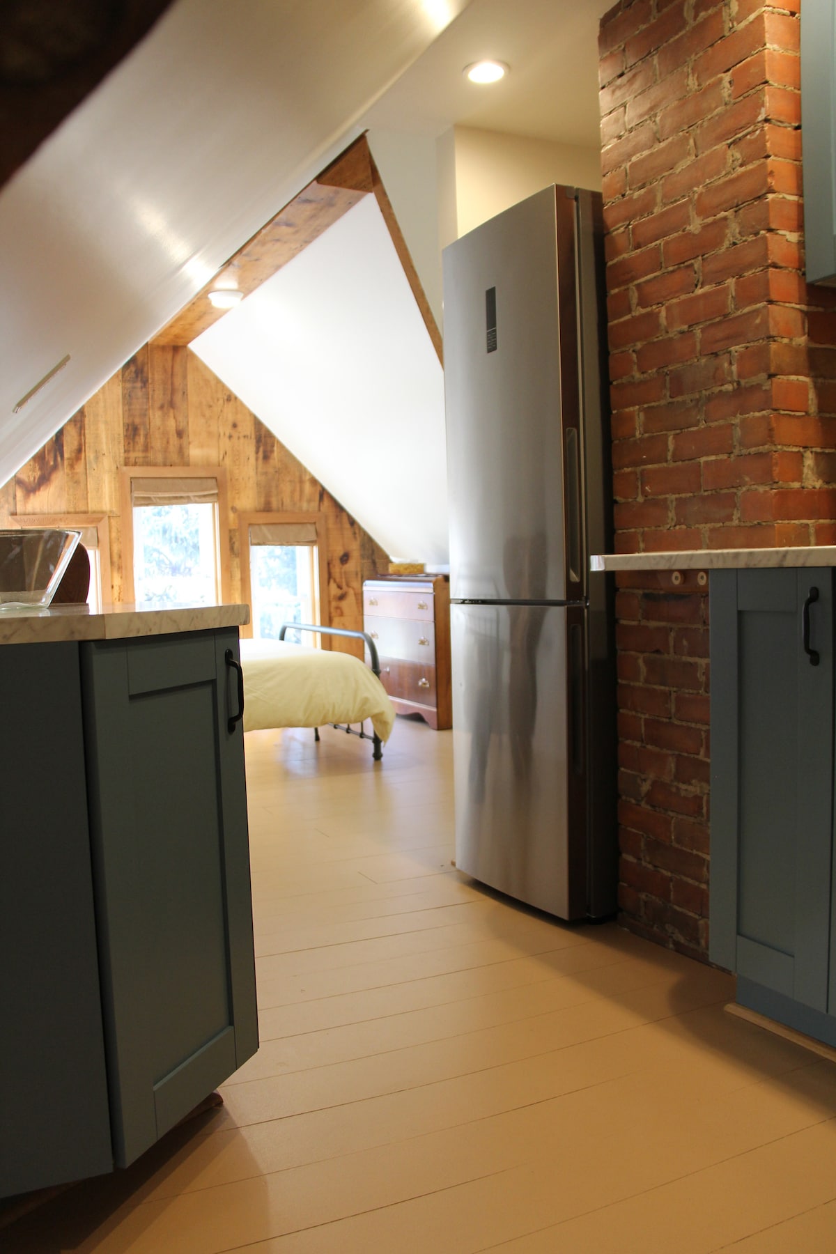 The kitchen area features sleek appliances, including a stainless steel refrigerator, set against a backdrop of rustic brick and wooden walls. Light floods in from the windows, illuminating the spacious layout and highlighting the soft tones of the cabinetry.