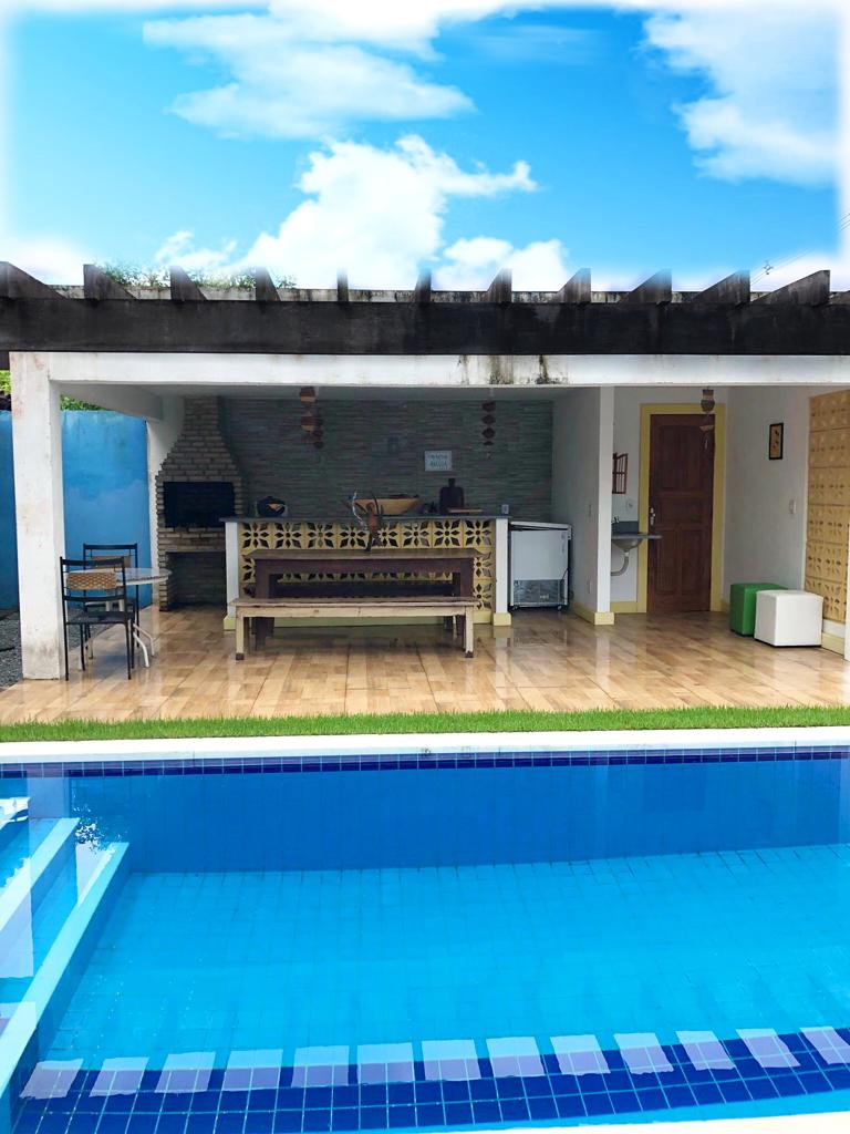 An outdoor gourmet area is situated next to a pool, featuring a barbecue grill and a counter for food preparation. A dining table with chairs is positioned nearby. The space is sheltered by a pergola, and clear skies can be seen above.
