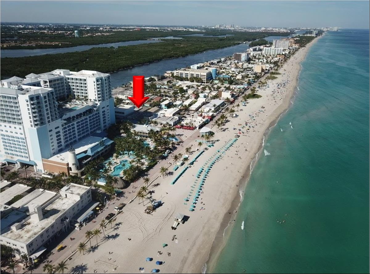 An aerial view captures the vibrant Hollywood Beach shoreline, highlighting the sandy beach and clear blue ocean. Lush greenery is visible in the background, along with nearby buildings. The location of the property is indicated with a red arrow, showcasing its proximity to the lively Broadwalk.