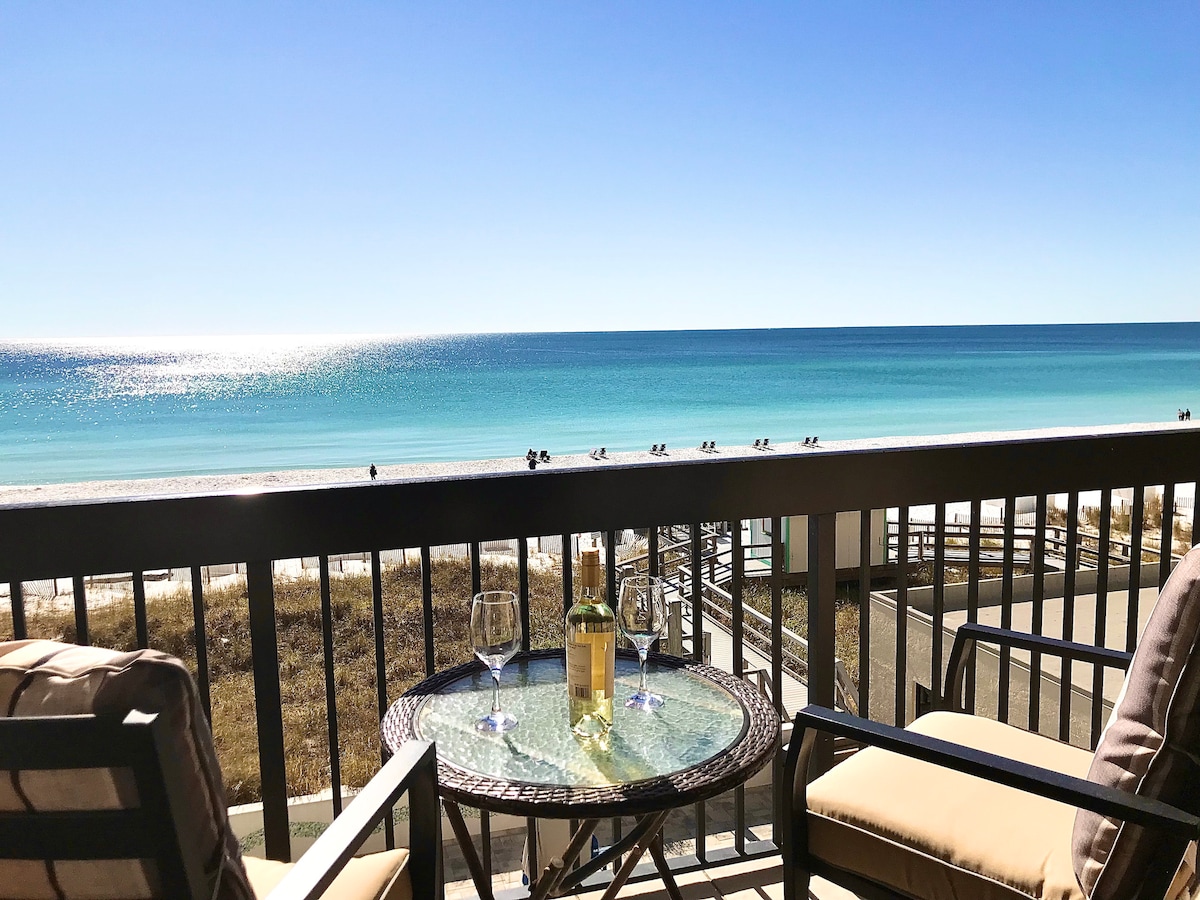 A balcony view presents the Gulf of Mexico, showcasing a vibrant blue horizon. A glass table is surrounded by two chairs, with two glasses resting on the table. The sandy beach is visible in the foreground, enhancing the serene coastal atmosphere.