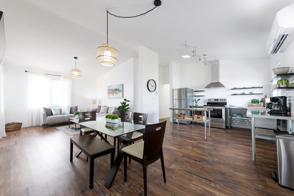 An open-plan living area features a dining table with dark wood chairs, complemented by a modern kitchen equipped with stainless steel appliances. Natural light filters through sheer curtains, enhancing the warm wood flooring. A comfortable sofa is positioned in the corner, creating a relaxed living space.