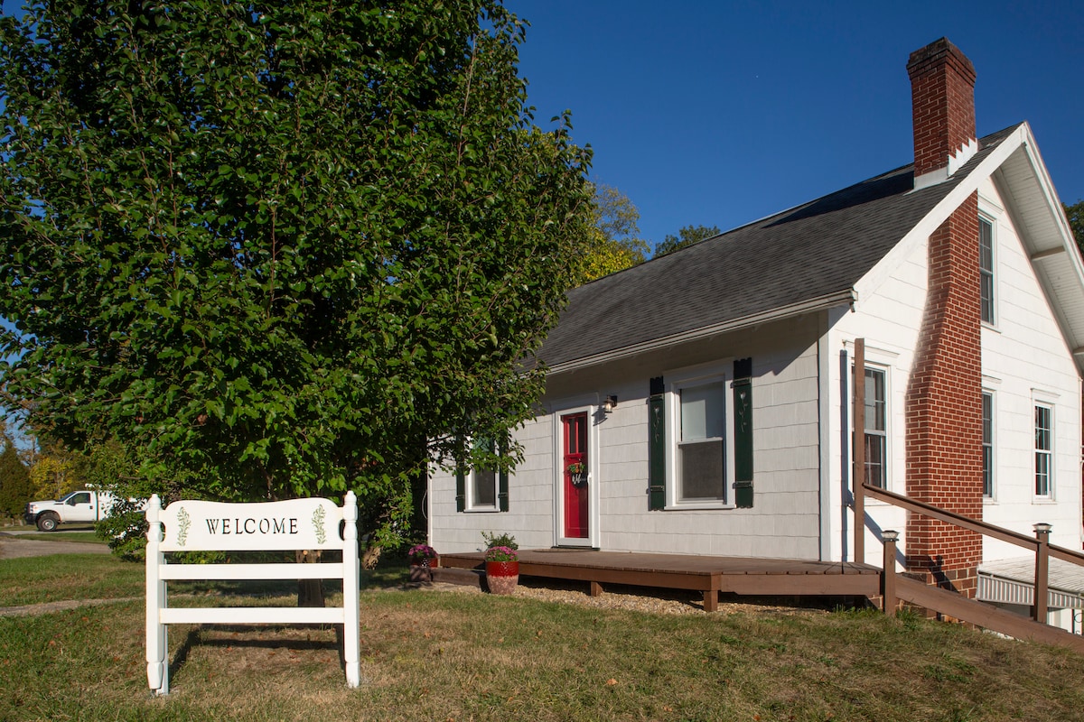 The exterior of a charming cottage is depicted, featuring white siding and a red front door. A 'Welcome' sign is displayed on a wooden stand in the foreground, complemented by a well-maintained yard with a large tree and a small flower pot.