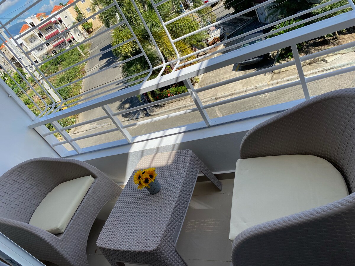 A balcony is featured, furnished with two woven chairs and a small table. A vase with sunflowers sits on the table. Sunlight illuminates the space, creating a bright ambiance. The view includes passing vehicles and greenery along the street below.