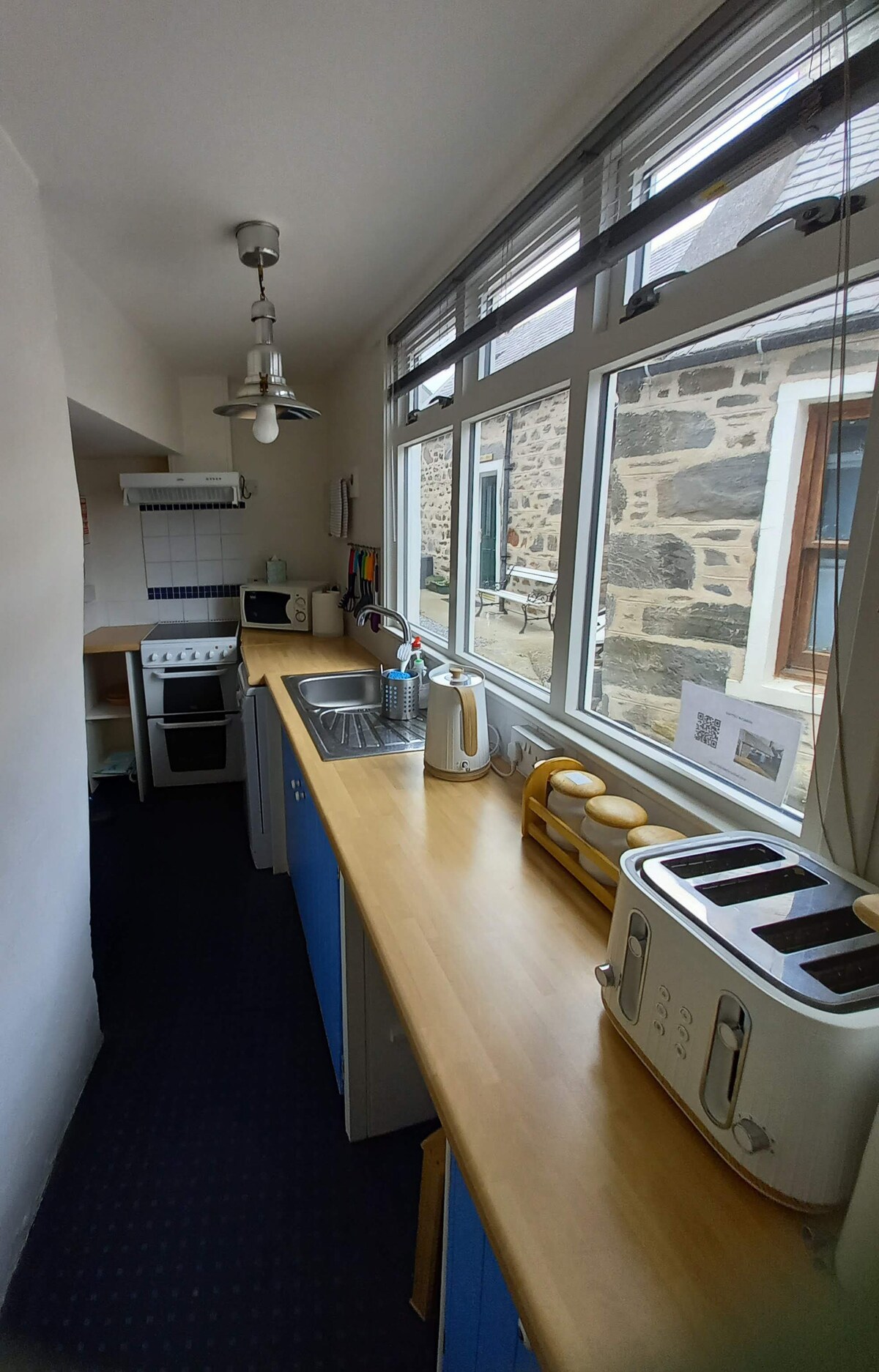 A narrow kitchen is fitted with modern appliances, including a stove, microwave, and a toaster. A wooden countertop runs along one side, with a sink and bright natural light filtering in through large windows. Stone walls add texture to the inviting space.