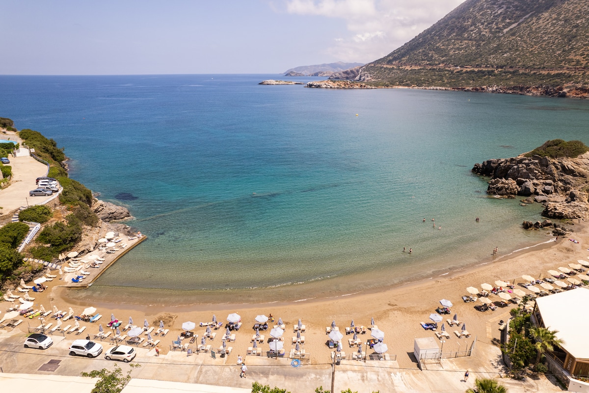 A serene beach scene presents soft golden sand meeting calm turquoise waters. Sun loungers and umbrellas line the shore, while gentle waves lap at the beach. In the background, a mountainous landscape rises, framing the peaceful coastal view.