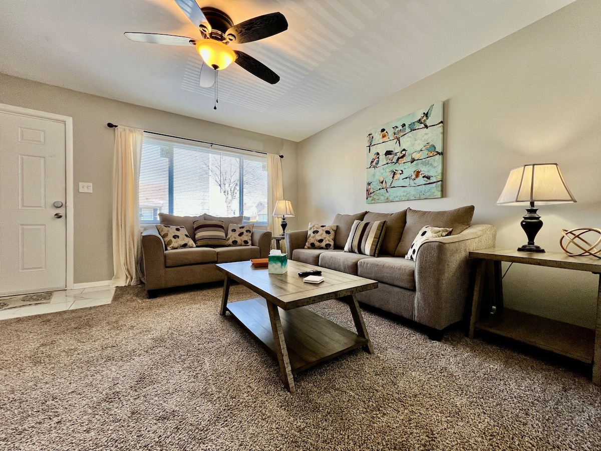 The living room features a welcoming layout with a spacious sofa and loveseat, complemented by a coffee table at the center. A ceiling fan is present, and natural light enters through a large window, enhancing the comfortable ambiance. Decorative artwork adds a touch of character.