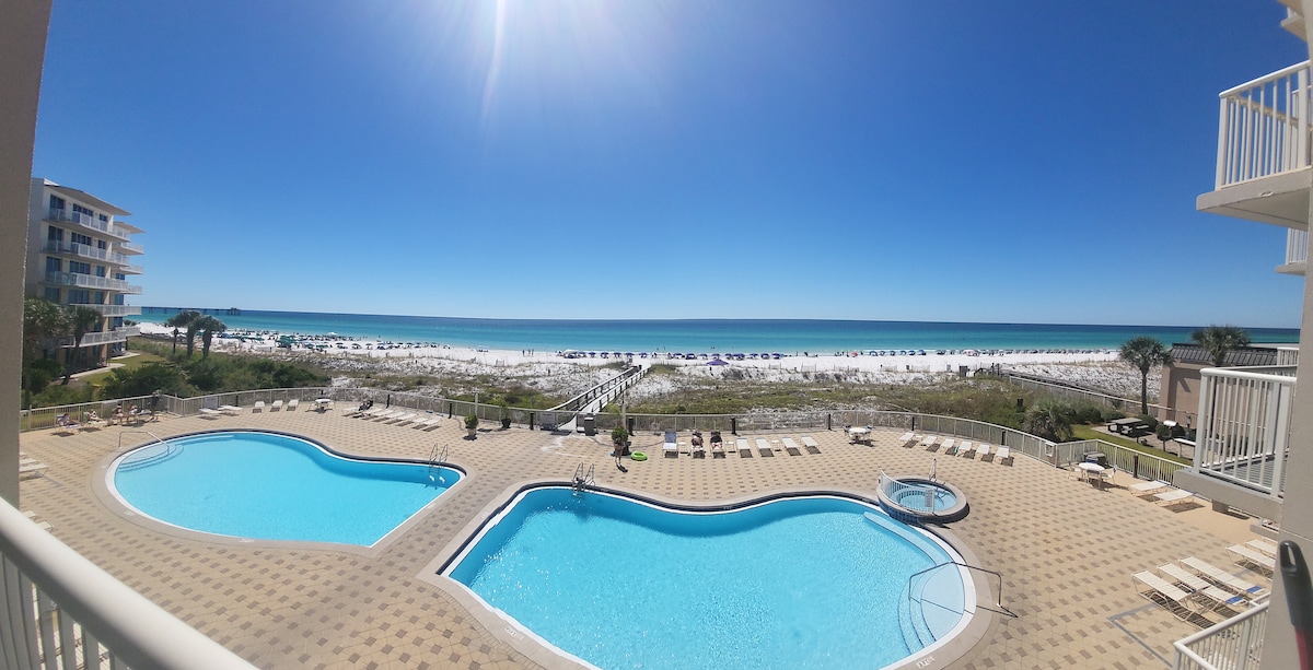 Two beach-side pools are visible next to a wide expanse of white sandy beach leading to the emerald waters of the Gulf Coast. Clear skies dominate the scene, while rows of beach chairs are positioned along the shoreline.