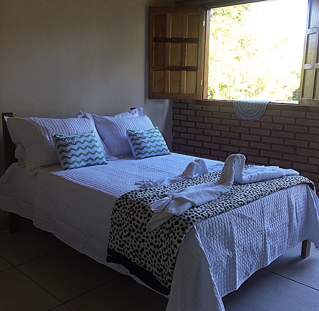 A bright sleeping area features a spacious bed adorned with a white quilt, patterned throw, and decorative cushions. Natural light floods the room through a window with wooden shutters, revealing a glimpse of the surrounding greenery outside.