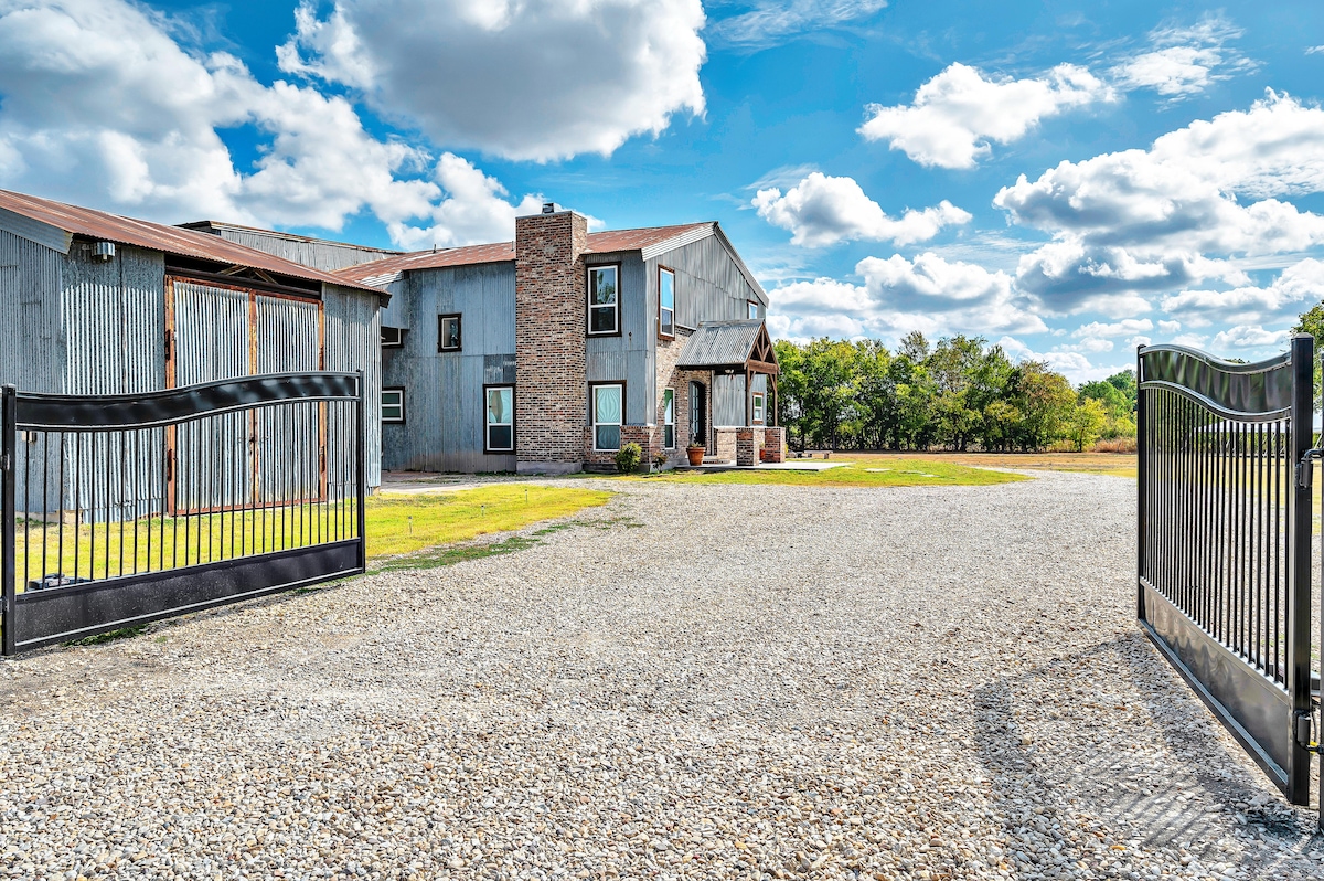 An expansive gravel driveway leads to a rustic home featuring a blend of corrugated metal and stone accents. A large gate opens to the property, framed by a lush green landscape under a bright blue sky dotted with fluffy white clouds.
