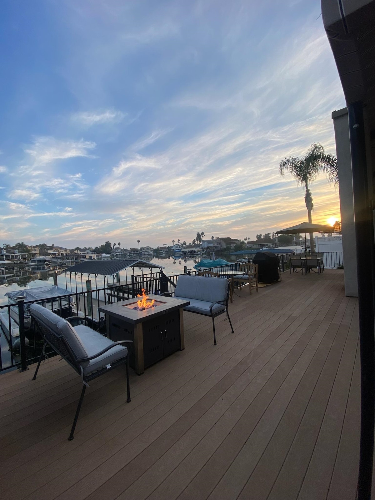 An outdoor deck features a cozy seating arrangement with a fire pit at the center. Water views extend into the distance, complemented by a sunset sky. Comfortable chairs are positioned around the fire, creating an inviting atmosphere for relaxation.