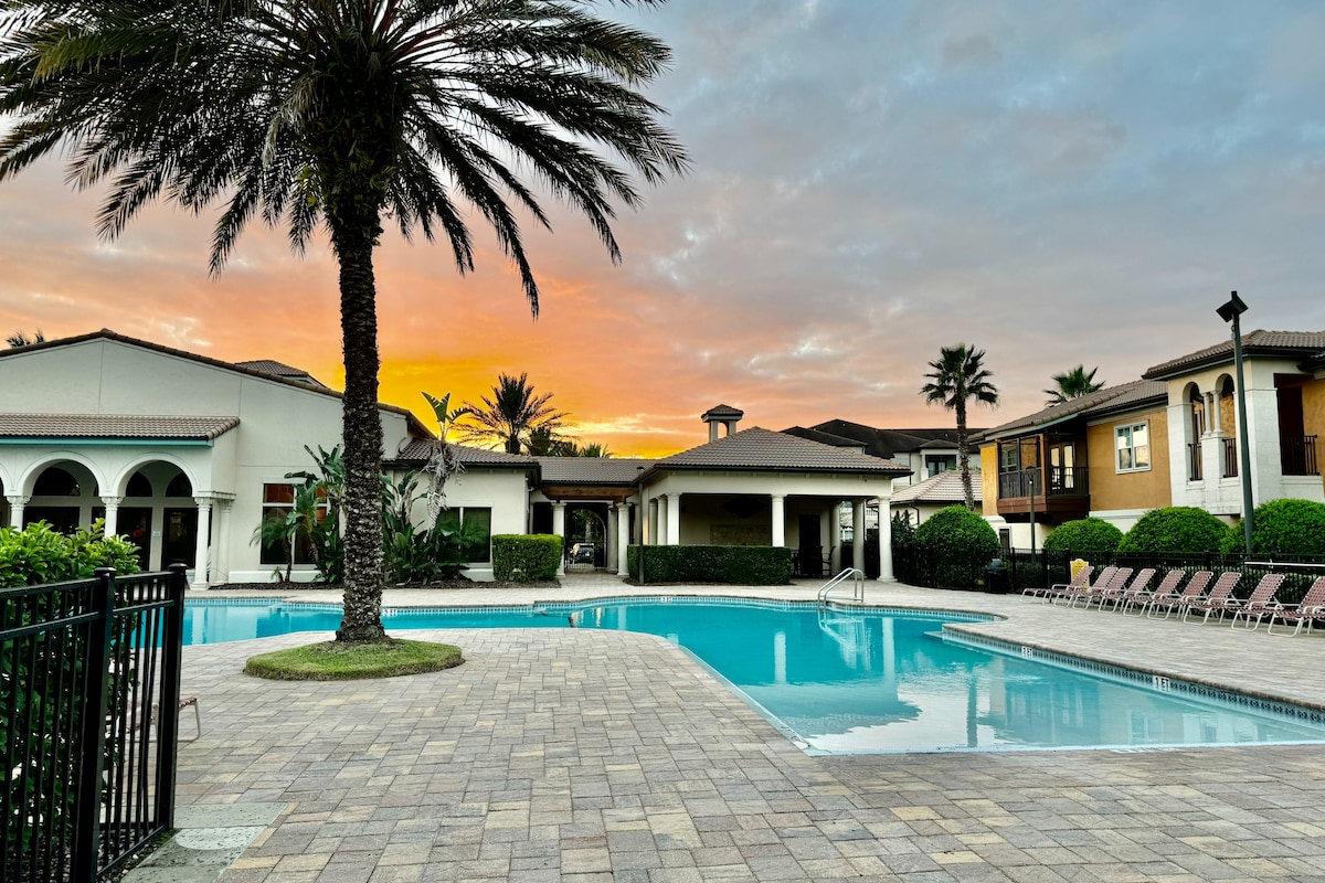 A serene pool area is presented, surrounded by palm trees and neatly landscaped greenery. The pool reflects the warm hues of an early morning sky, with lounge chairs arranged along the poolside. The buildings of the resort-style complex are visible in the backdrop.