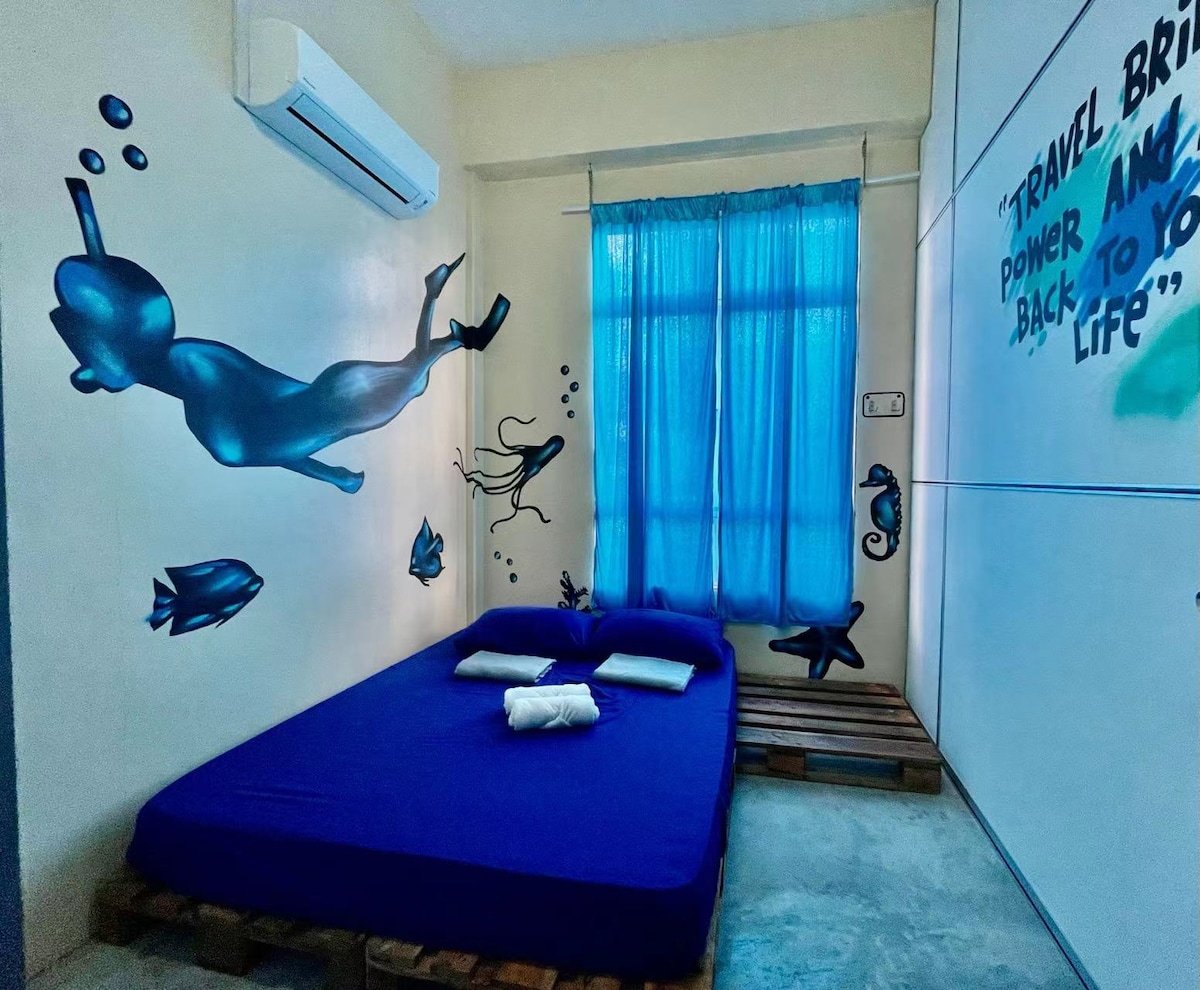 A unique bedroom features a purple mattress on a wooden pallet with three white pillows arranged on it. Bright blue curtains frame a window, while colorful wall art depicting underwater scenes adds character to the space.