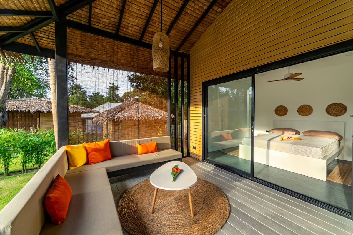 A covered terrace lounge area features a sectional sofa with colorful cushions and a round coffee table. Large glass doors provide a view of the inviting sleeping area inside, designed with a spacious bed and natural decor accents. Surrounding greenery and thatched-roof bungalows are visible in the background.