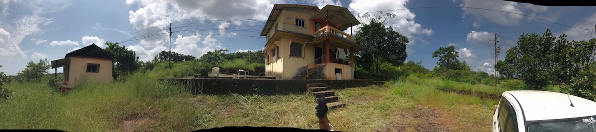 A two-story house is viewed from the front, featuring a wide staircase leading to a spacious porch. Surrounding greenery adds to the natural setting, with open outdoor areas indicated for relaxation. A smaller structure is seen to the left, further enhancing the tranquil environment.