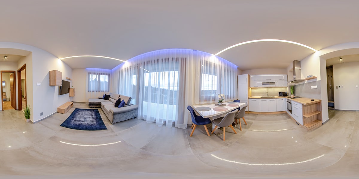 A spacious living area is visible, featuring a comfortable sectional sofa and a dining table surrounded by stylish chairs. Soft lighting illuminates the room, highlighting the modern kitchen with wood accents. Large windows offer ample natural light and views of the surroundings.