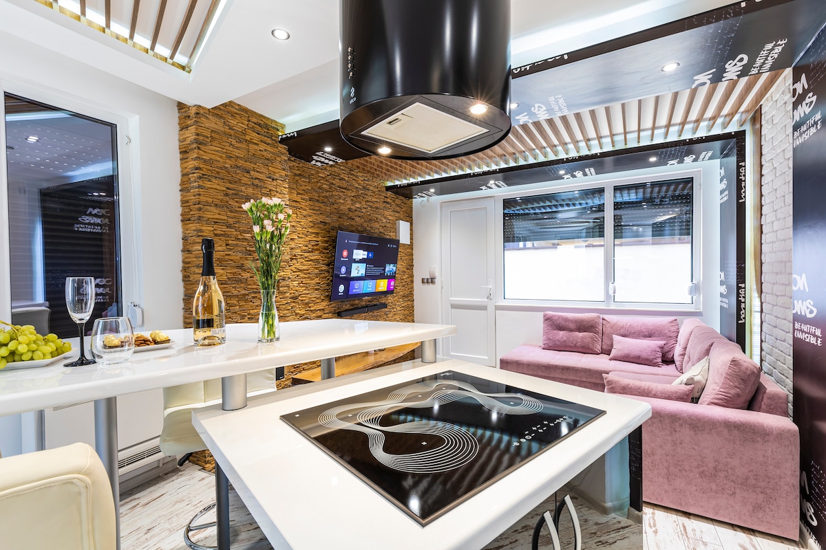 The modern living space features a stylish open-plan layout with a comfortable sofa and a flat-screen TV mounted on a feature wall. A central dining table is complemented by an elegant kitchen island, while large windows offer ample natural light.