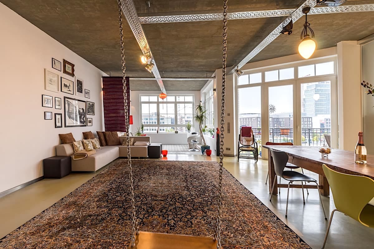A spacious living area features a comfortable couch and a decorative rug, creating a relaxed atmosphere. A stylish swing hangs from the ceiling, adding a playful element. Large windows provide ample natural light and a view of the vibrant outdoor space, while a contemporary dining table sets the scene for shared meals.