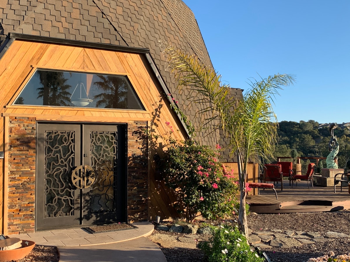 A unique entrance is highlighted by an ornate black door with decorative ironwork. Surrounding flora adds color, while a sculpture and outdoor seating area are visible in the background, along with the natural landscape and clear blue sky.