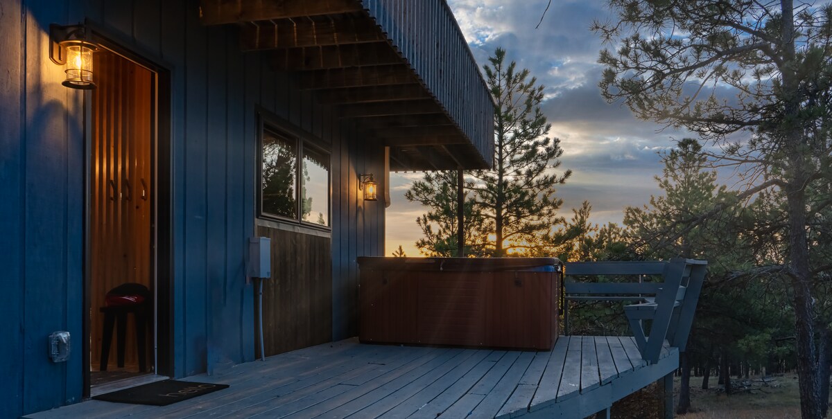 A spacious deck extends from the building, featuring a hot tub under warm evening light. Large windows provide views of the surrounding trees, while two outdoor lights illuminate the entryway. A stool is positioned near the door, enhancing convenience.