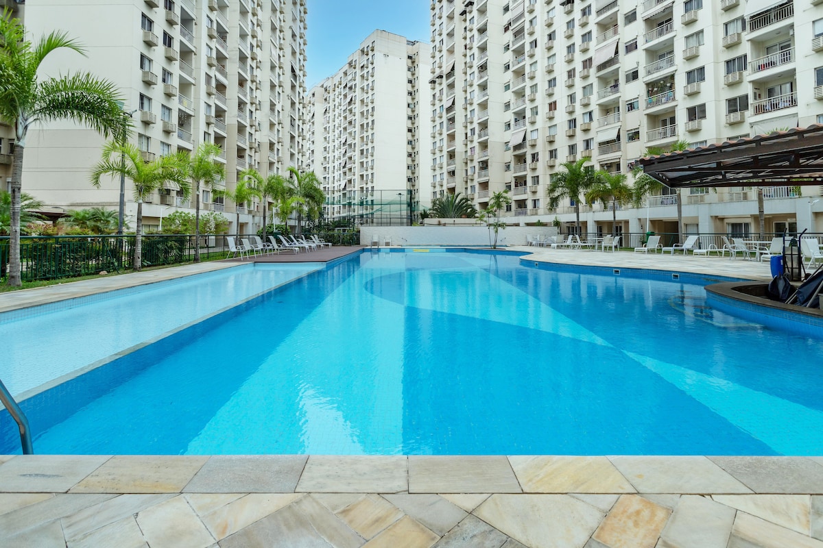 A large swimming pool is featured at the center of the condominium complex, surrounded by tall buildings. Lush palm trees are positioned along the pool's edge, with numerous lounge chairs available for relaxation. The water appears clear and inviting under a bright blue sky.