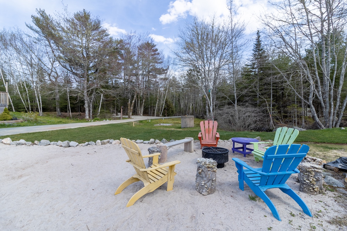 A sandy area features four colorful Adirondack chairs arranged around a small fire pit. Stone borders outline the seating area, with a backdrop of trees providing natural privacy and tranquility. The scene invites relaxation and enjoyment of the outdoor space.