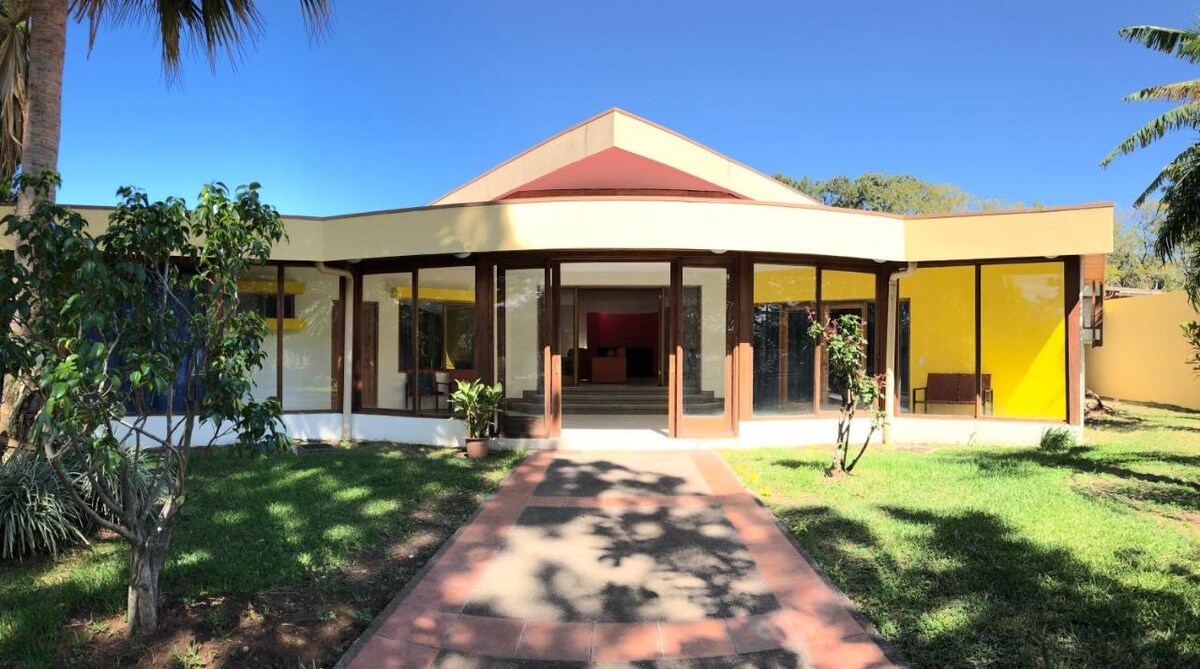 A modern one-story structure features a bright yellow accent wall and large glass windows, creating an airy atmosphere. The path leading to the entrance is bordered by greenery, contributing to a welcoming exterior. Palm trees and flowering plants frame the building, enhancing the outdoor environment.