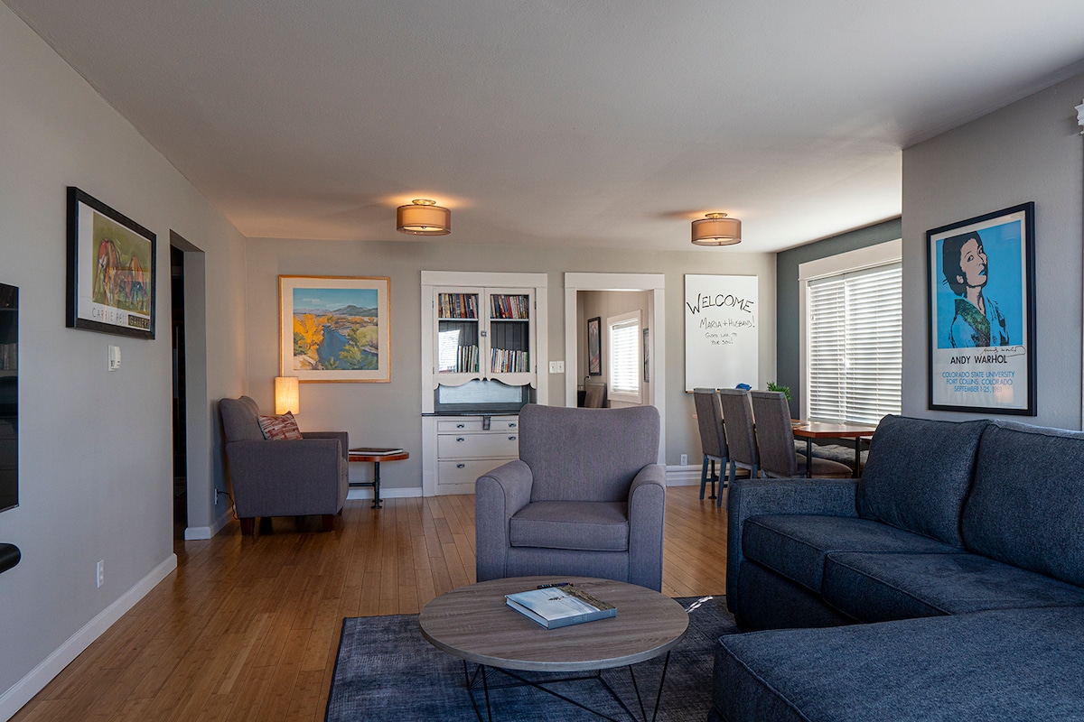 A spacious living area features a comfortable gray sofa and an accent chair, arranged around a round coffee table. Light wood floors complement the neutral-colored walls. Artistic prints decorate the space, which is well-lit by natural light streaming through the windows.
