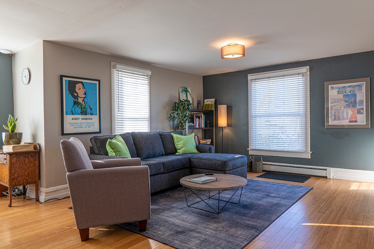 A modern living room features a sectional sofa with green accent pillows and a coffee table in the center. Large windows bring in natural light, while a cozy rug lays beneath. A side table and a plant add warmth to the space, complemented by framed artwork on the walls.