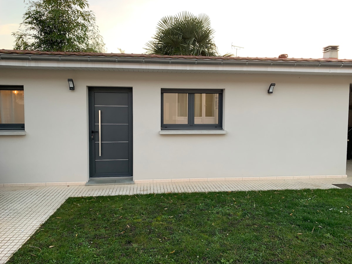 The exterior of a modern single-story building is depicted, featuring a sleek gray door with a horizontal handle. A large window is present, framed by light-colored walls and surrounded by a small well-maintained lawn. Outdoor lighting enhances the entryway, creating a welcoming appearance.