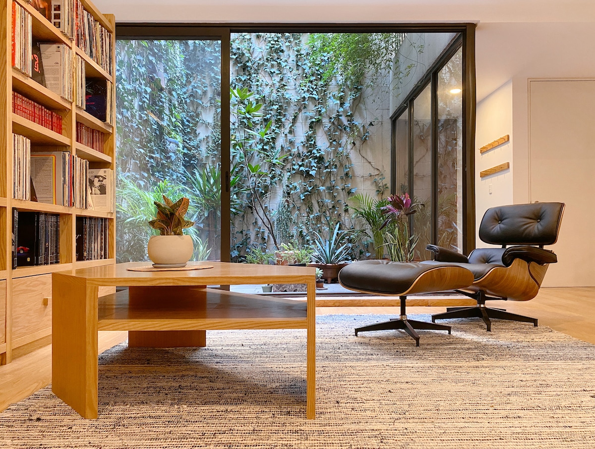 A cozy living space is highlighted by a large window overlooking a lush green terrace filled with plants. A classic Eames Lounge Chair and a wooden coffee table are positioned on a textured area rug, offering a comfortable spot to relax or unwind.