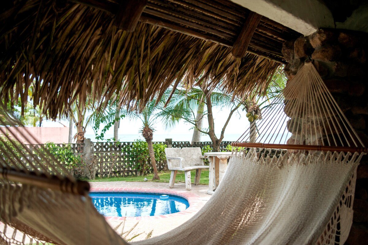 A thatched roof provides shade over a relaxing outdoor space. A hammock is suspended nearby, offering a view of a pool surrounded by greenery. A stone table and benches are positioned under palm trees, contributing to a serene ambiance.