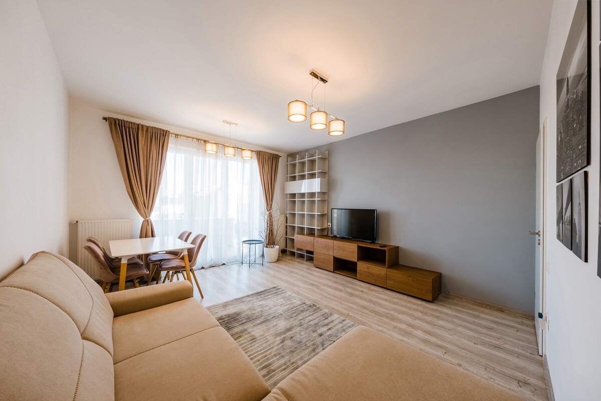 A spacious living room features an extendable sofa, a wooden TV stand, and a dining table with chairs. Large windows are dressed with soft curtains, allowing natural light to fill the area. Wood flooring adds warmth to the contemporary design.