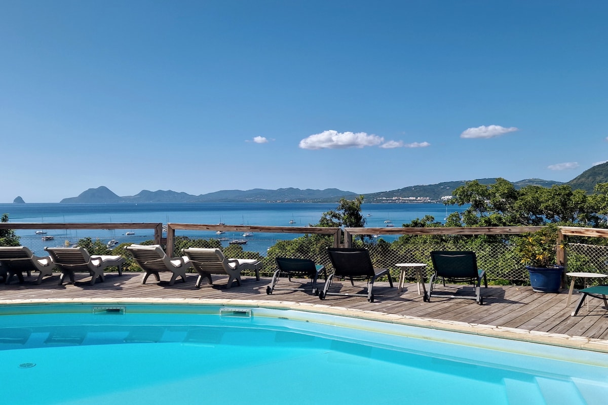 A shared swimming pool is visible in the foreground, surrounded by a wooden deck with multiple lounge chairs positioned for relaxation. Beyond, a sweeping view of the Caribbean Sea and distant islands can be seen under a clear blue sky.