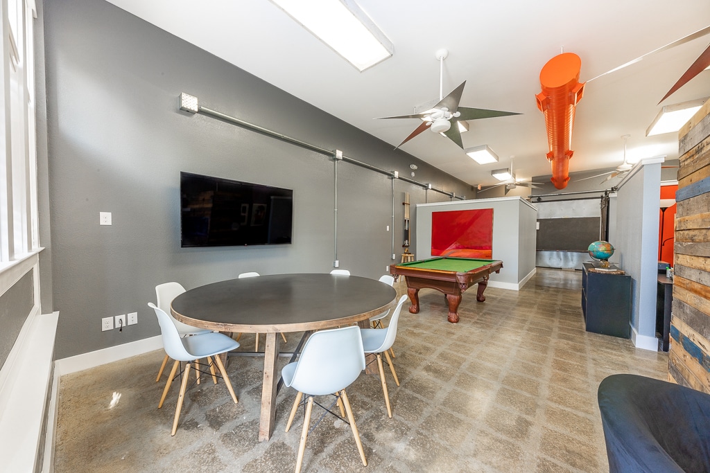 A spacious common area features a round dining table with white chairs, positioned under overhead lights. A wall-mounted television is visible, along with a full-size pool table in the background. Exposed ducts and a ceiling fan add to the industrial design.