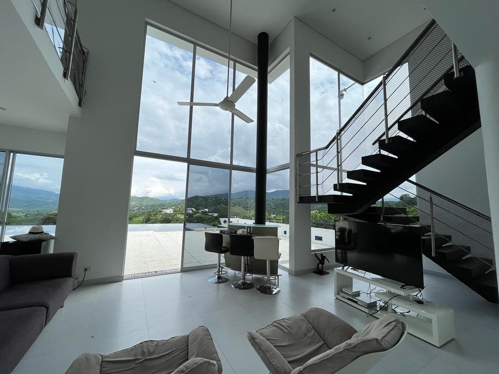 A bright and airy living area showcases expansive floor-to-ceiling windows, providing a view of the mountains and outdoor spaces. An elegant staircase leads to the upper level. Comfortable seating areas and a modern TV setup are positioned within this open-concept design.