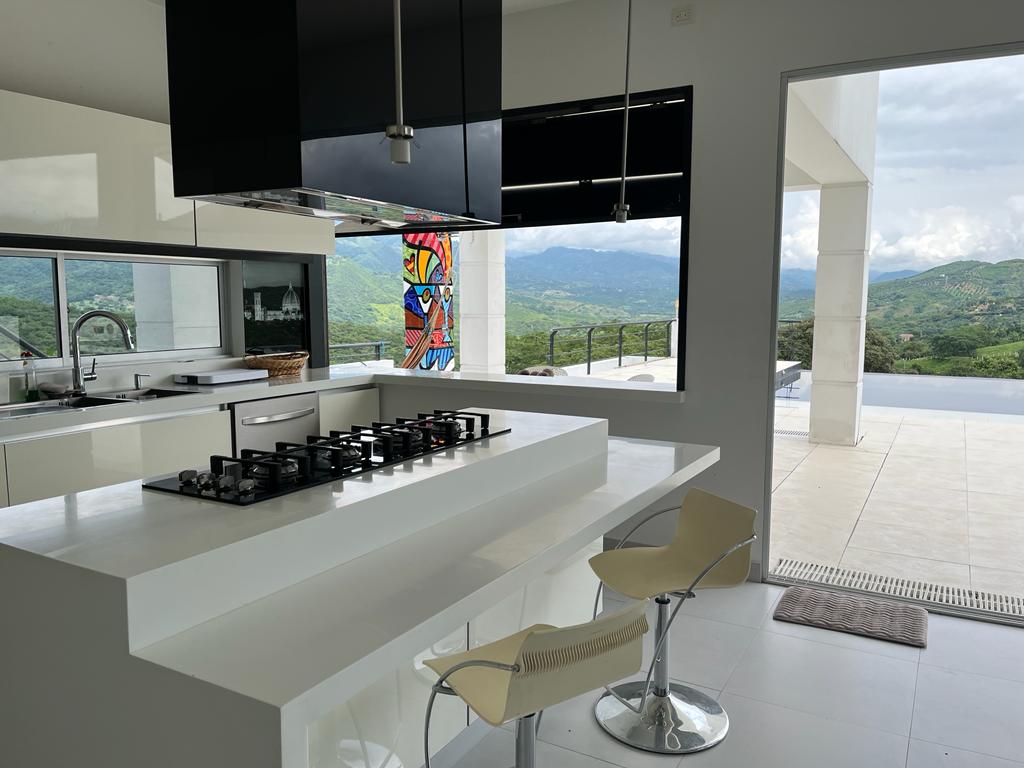 A modern kitchen features a minimalist design with a central island and a gas cooktop. Large windows provide a view of the surrounding landscape, while bright colors from an artwork enhance the space. Stylish seating is available at the island.