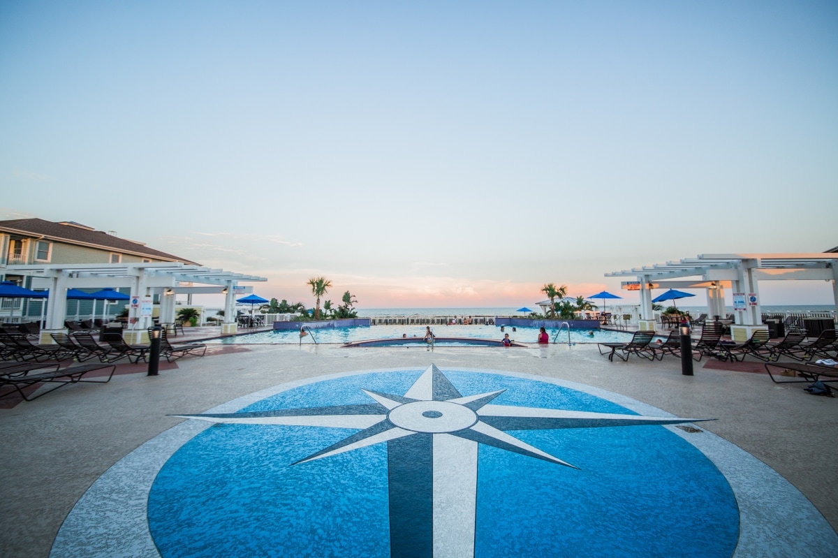 A resort pool area features a central compass design on the deck, surrounded by lounge chairs and umbrellas. The pool extends towards the horizon, where the sky meets the ocean, creating a serene backdrop. The atmosphere is enhanced by palm trees and a calm sunset.