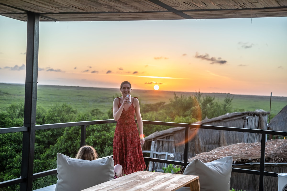 A scenic outdoor terrace showcases a sunset over the lush landscape, viewed from a comfortable seating area. A person stands on the balcony, enjoying a drink, while another sits casually in the background. Soft cushions are present, enhancing the relaxed atmosphere.