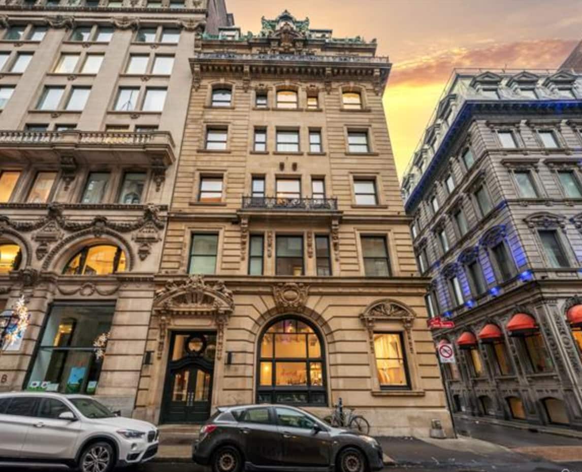 An elegant, historic building stands prominently on a bustling street. Its grand façade features ornate architectural details, large windows, and a welcoming entrance. Surrounding buildings reflect a blend of classic and modern styles, while a car is parked nearby, demonstrating the urban atmosphere of the area.