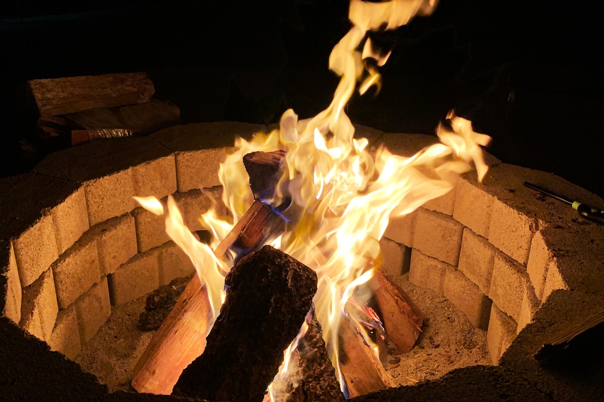 A circular stone fire pit is filled with bright flames and glowing embers. Logs are arranged within the pit, some partially consumed by fire, creating a warm glow against the dark night backdrop.