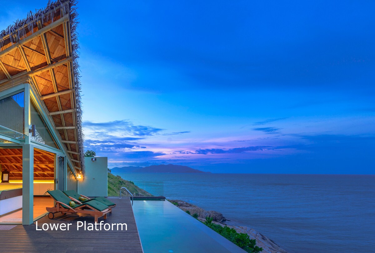 An oceanfront terrace is illuminated by early evening light, featuring lounge chairs positioned for scenic views. The infinity pool extends towards the horizon, reflecting the soft colors of the sunset over the sea. The architecture includes a bamboo roof that complements the natural surroundings.