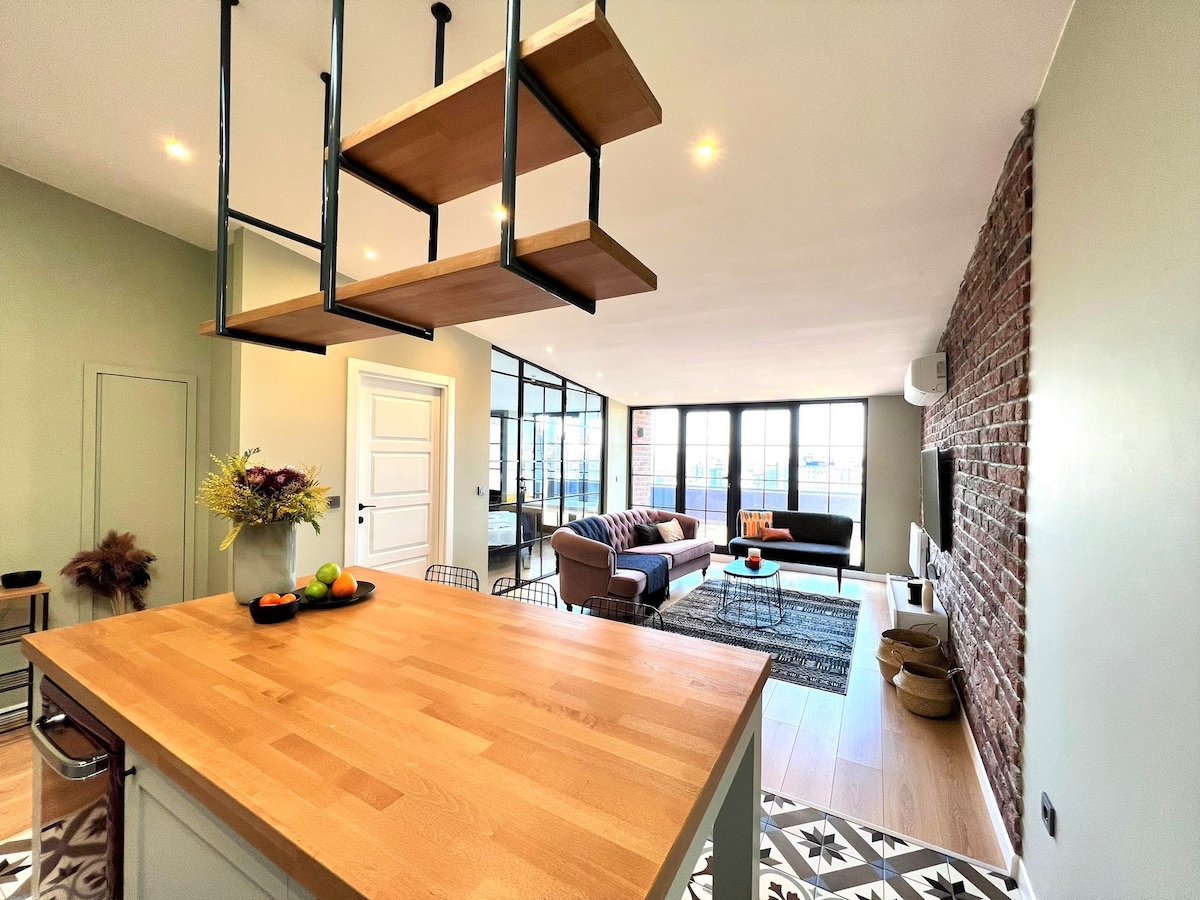 The open-plan living space features a wooden kitchen island illuminated by suspended shelves, with fresh fruit displayed. A cozy seating area is arranged with a sectional sofa and decorative rug, while large windows allow ample natural light to fill the room.