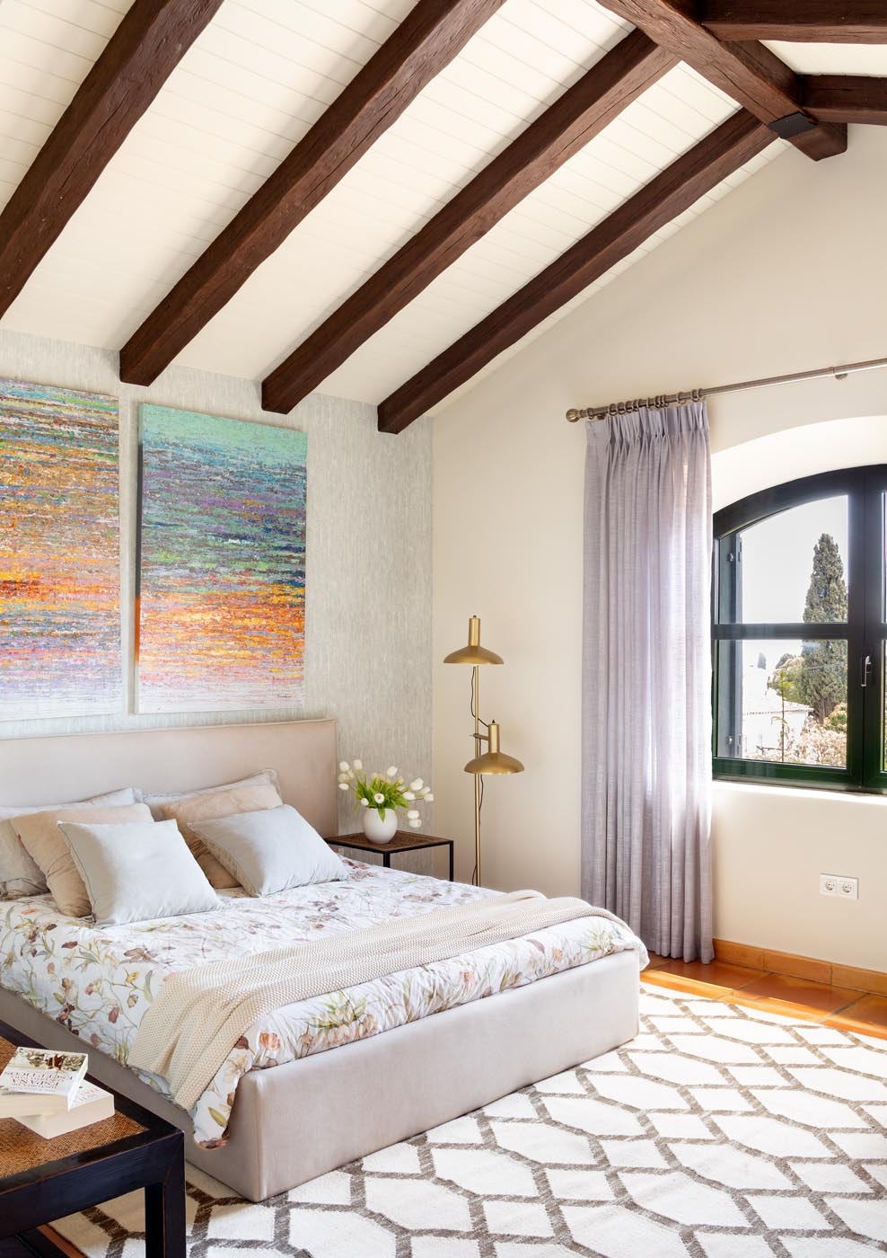 A serene bedroom features a spacious bed adorned with a floral comforter. High, wooden beams frame the ceiling, and two vibrant art pieces hang above the bed. Natural light filters through a large arched window, complemented by soft curtains.