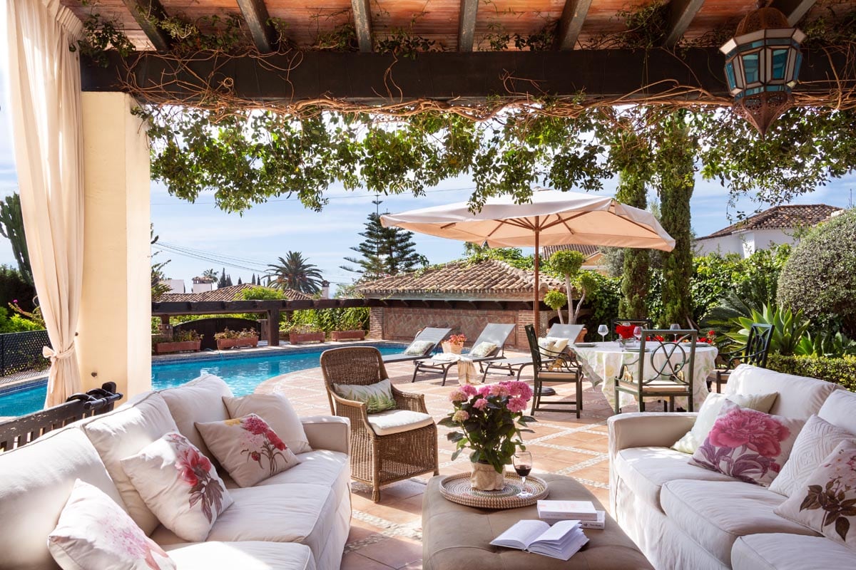 A tranquil outdoor area features a spacious seating arrangement with soft cushions, set beneath a pergola adorned with climbing vines. A large pool is visible in the background, surrounded by sun loungers and lush greenery, offering a serene environment for relaxation.