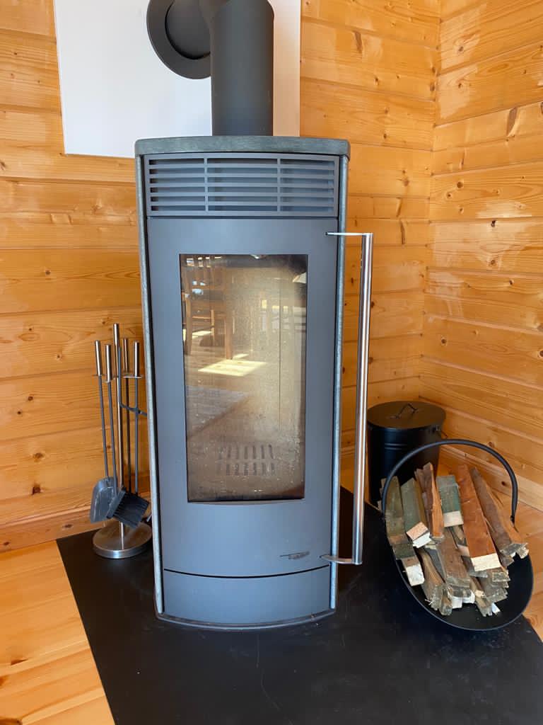 A modern wood stove, positioned in the corner of a wooden cabin, features a large glass front that showcases a view of the flames inside. Nearby, a basket holds neatly arranged logs, accompanied by set tools for maintenance.