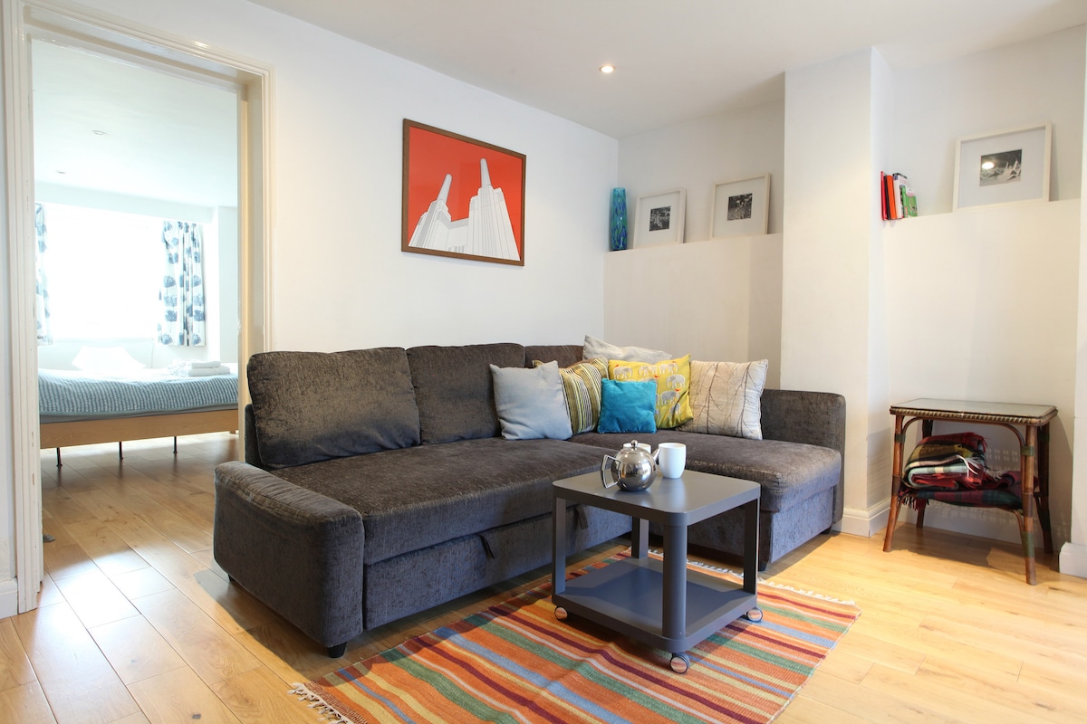 A spacious living room area features a dark grey sectional sofa adorned with colorful cushions. A low coffee table sits in front, accompanied by a striped area rug. Wall art adds a pop of color, while natural light pours in through large windows in the background.