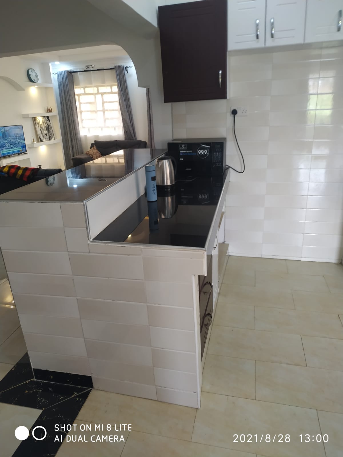 The kitchen area is equipped with modern appliances, including a kettle and microwave. A sleek black countertop offers ample space for meal preparation. Light-colored tiles contribute to a clean and bright atmosphere, while a window allows natural light to enhance the space.