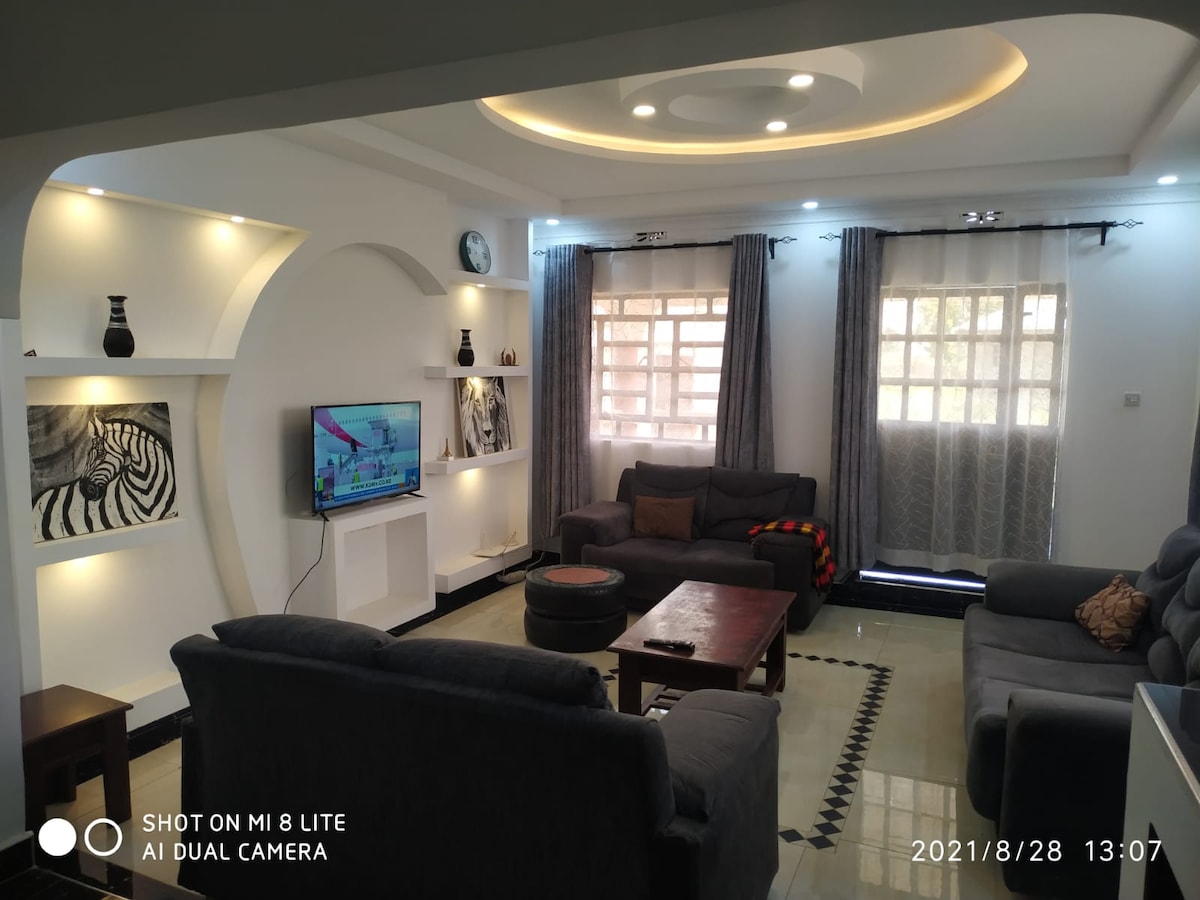 A spacious living area is illuminated by soft, recessed lighting. A large television is centered on a feature wall, complemented by artistic decor. Comfortable seating is provided by two gray sofas and an armchair, while natural light filters through sheer curtains at the windows.