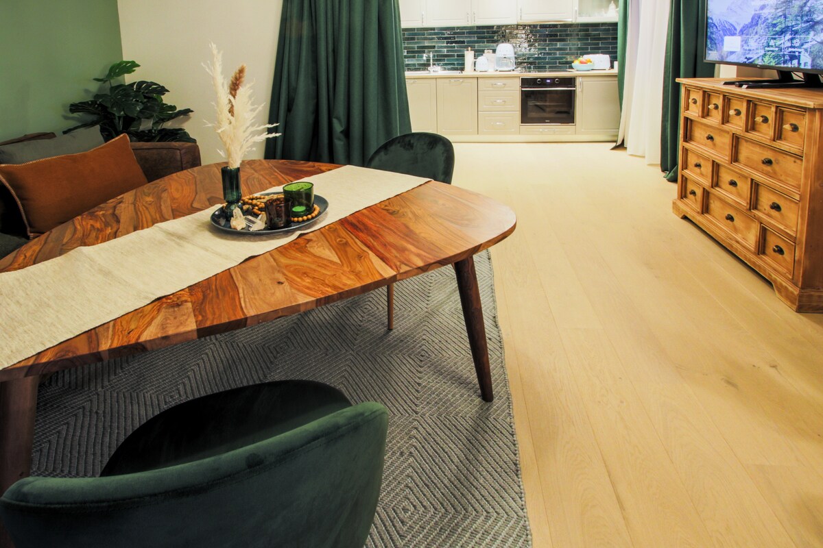 A wooden dining table with a detailed surface is complemented by plush green chairs. A crafted centerpiece sits on the table, surrounded by a decorative runner. In the background, a well-equipped kitchen is visible, along with a wooden sideboard and a television.